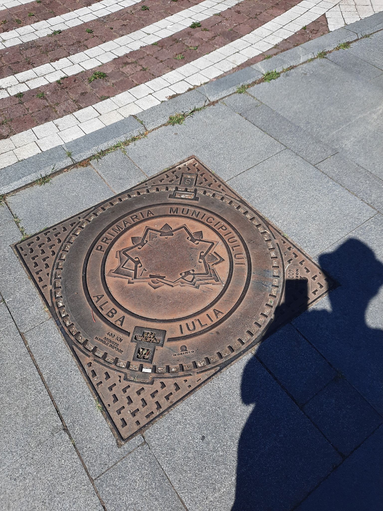 Gullideckel mit der Aufschrift Primaria Municipiukui Alba Iulia. In der Mitte ist die Silhouette der Festung Alba Iulia. Im Vordergrund ist Silkes Schatten, die das Handy in der Hand hält und dieses Foto macht.