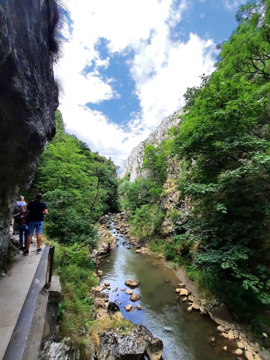 Ein Fluss fließt durch eine Schlucht mit grauen, von Büschen und Gras bewachsenen Felsen. Im Vordergrund stehen Bäume am Flussufer, dass voller großer Felsbrocken ist. Am linken Bildrand ist ein in den Fels gehauener betonierter Pfad, auf dem Menschen laufen