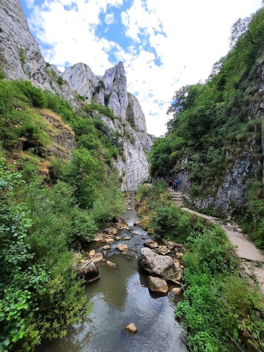 Ein Fluss fließt am Fuß grauer, von Büschen und Gras bewachsenen Felsen. Im Bildhintergrund sieht man eine Felsspitze, die einige Meter über dem restlichen Fels gen Himmel ragt. Rechts ist ein schmaler Weg, im Hintergrund laufen Menschen, zu klein um sie zu erkennen