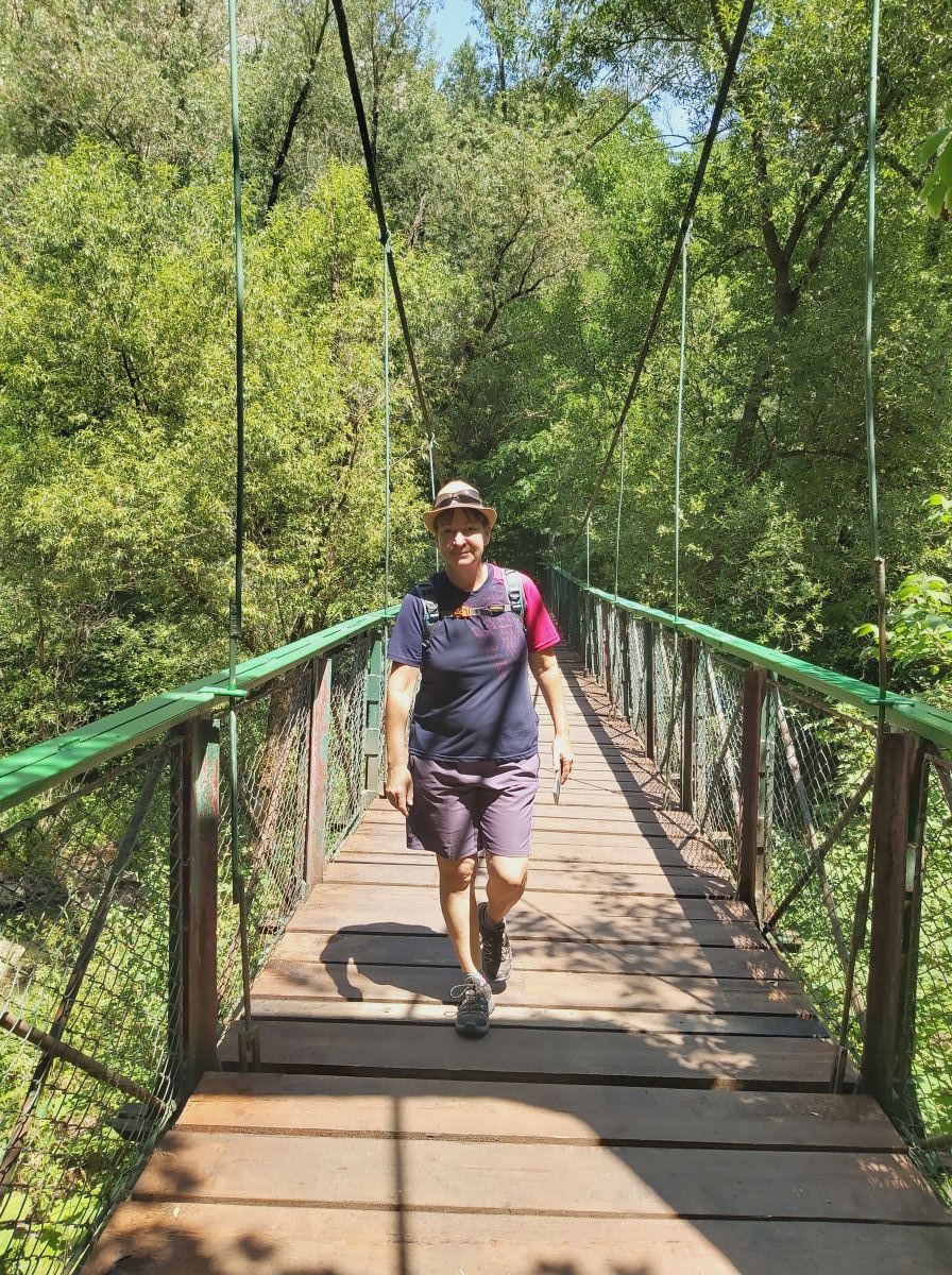 Silke läuft auf einer Brücke auf die Kamera zu. Sie trägt kurze hellviolette Hosen, ein blaues T-Shirt dessen rechter Aermel rot ist und einen beigen Strohhut. Die Brücke hat hölzerne Planken und ein mit grün lackiertes Geländer.