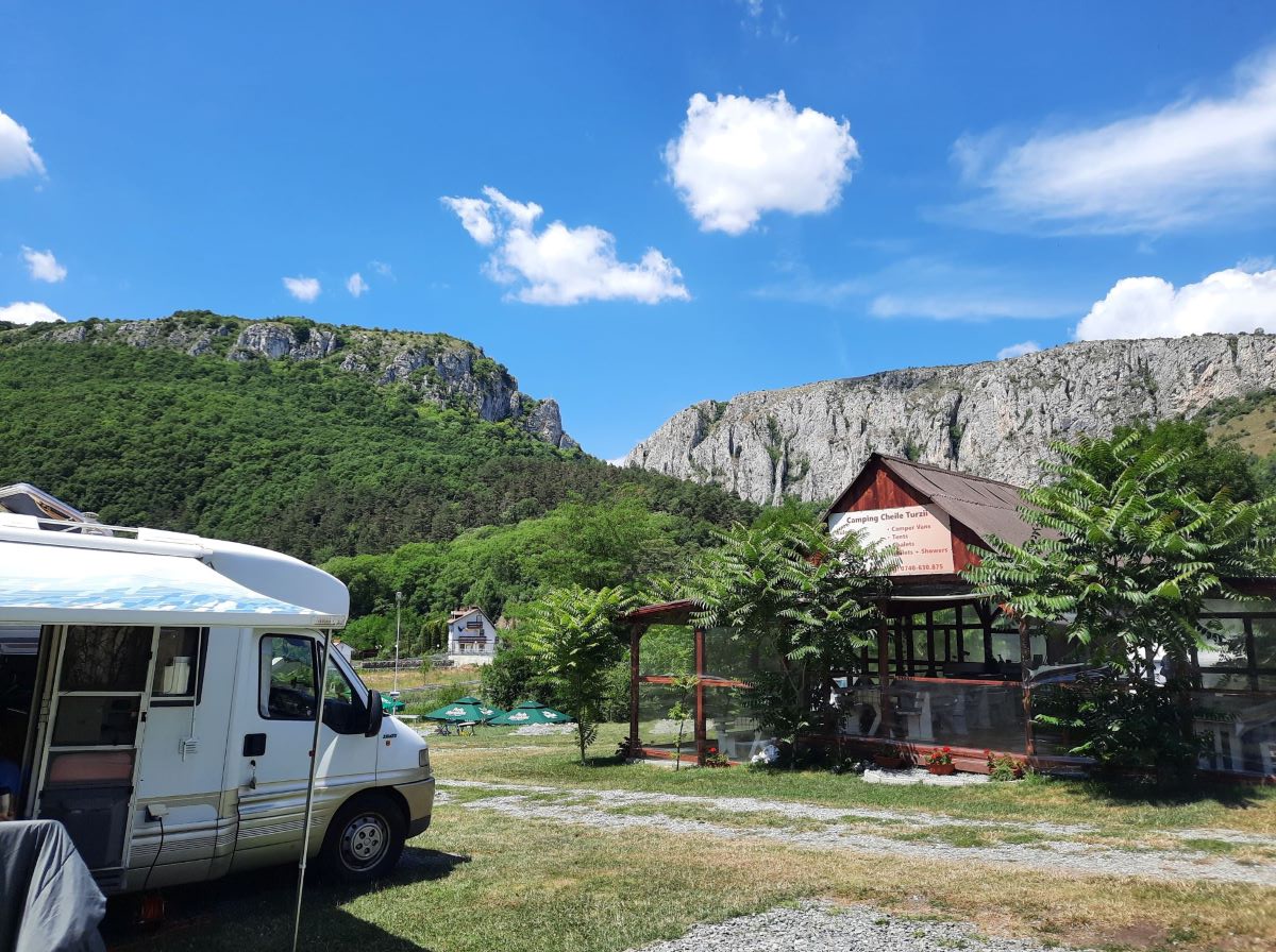 auf einer grünen Wiese steht am rechten Bildrand ein weißes Wohnmobil mit ausgefahrener Markise. Gegeüber steht ein hölzerner Pavillon. Im Hintergrund, inmitten von grünem Wald sieht man zwei steile graue Felswände, die in der Mitte von einer Schlucht getrennt sind. 