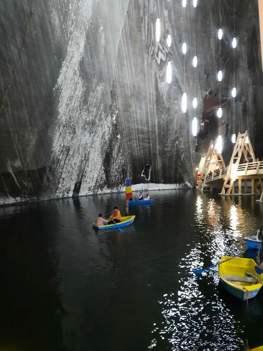 Im Vordergrund fahren kleine Ruderboote auf einem See im Tereza-Stollen der Salina Turda in Rumaenien. am rechten Bildrand eine hoelzerne Bruecke. Ueber dem Wasser haengen an langen Seilen mehrere schmale, hell leuchtende Lampen. Die steinerne graue Wand im Hintergrund ist von weißen gerade und unregelmaeßigen Linien durchzogen