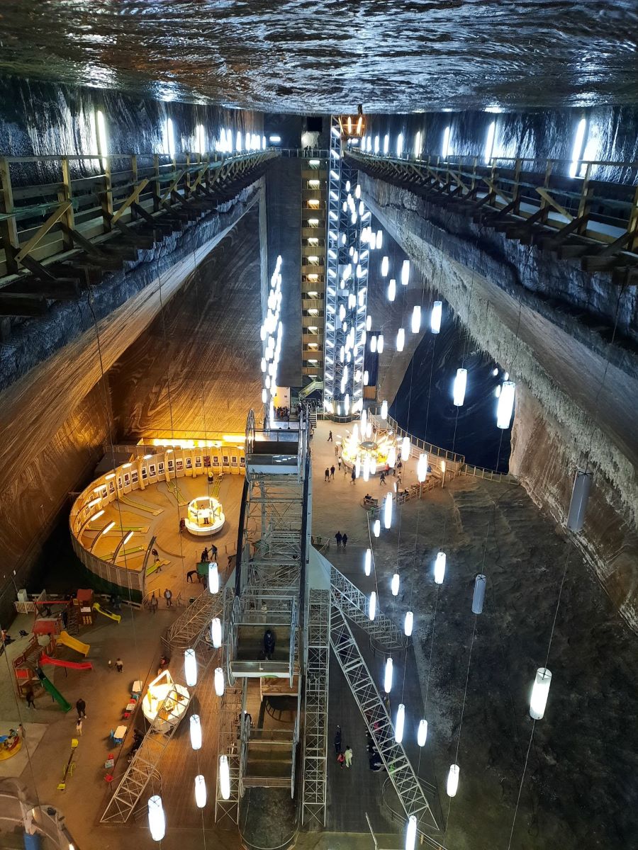 Blick von der Empore der Salina Turda in Rumaenien. Der Raum geht tief in das innere der Erde. Beleuchtet wird er von vielen schmalen, laenglichen Lampen, die an Seilen von der Decke haengen. In der Bildmitte sieht mand die Stahlkonstruktion eines Riesenrads und einen halbrund abgegrenzten Minigolf-Bereich. Am linken Bildrand sind mehrere Rutschen in gelb, rot und gruen zu sehen 