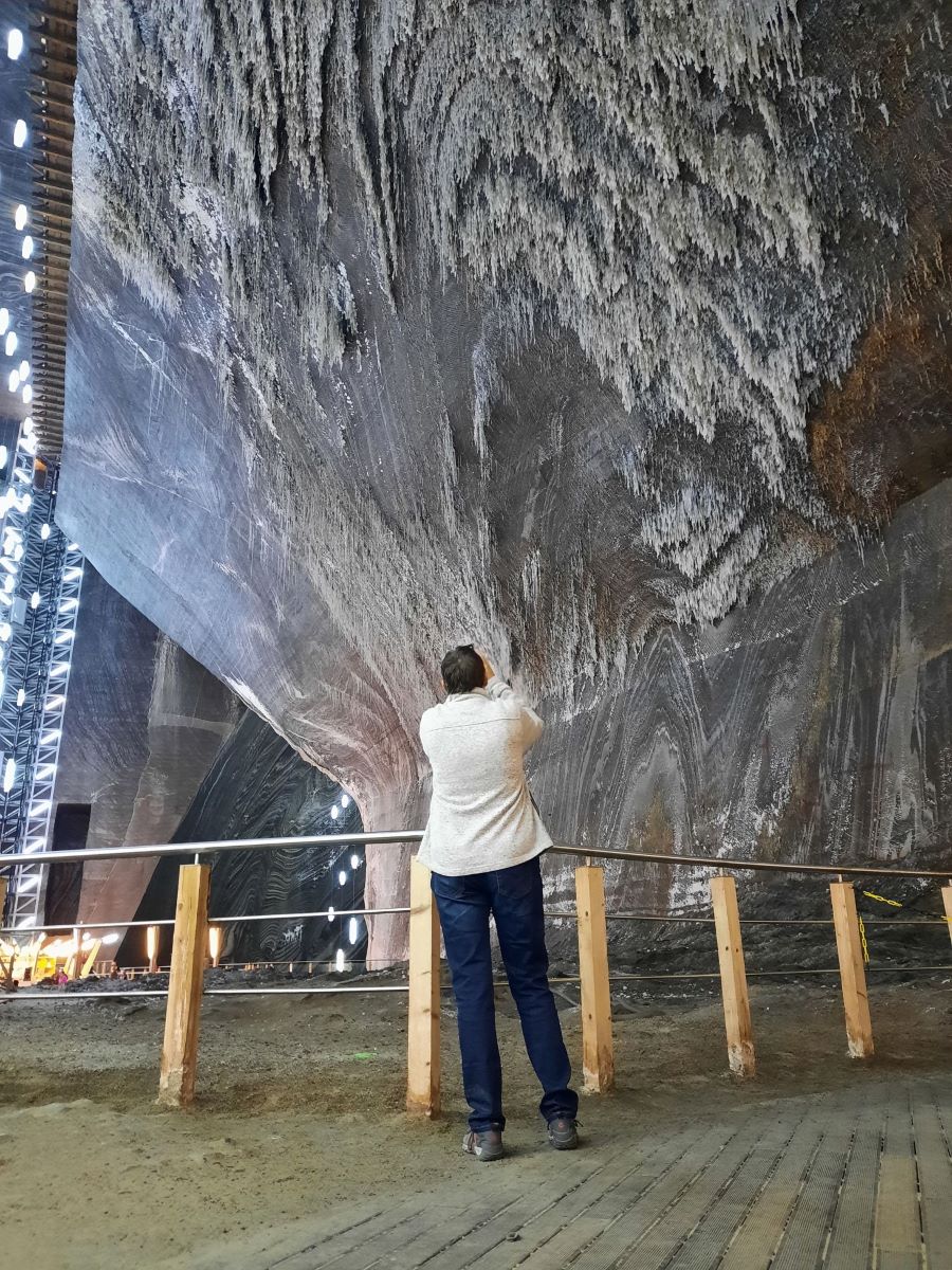 im Vordergrund steht Claudia mit ihrer grauen Jacke und blauen Jeans und fotografiert die schraege Decke der Salzmine Turda in Rumaenien. Salz kristallisiert in schmalen Stalagtiten von der Decke