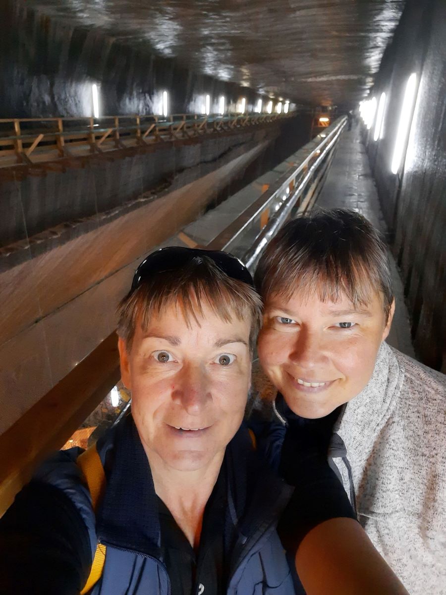 Im Vordergrund schauen Silke und Claudia in die Kamera. Silke traegt ein blaues Oberteil, Claudia eine grau gepunktete Jacke. Sie stehen auf der Empore der Salzmine Turda in Rumaenien, die im Hintergrund zu sehen ist