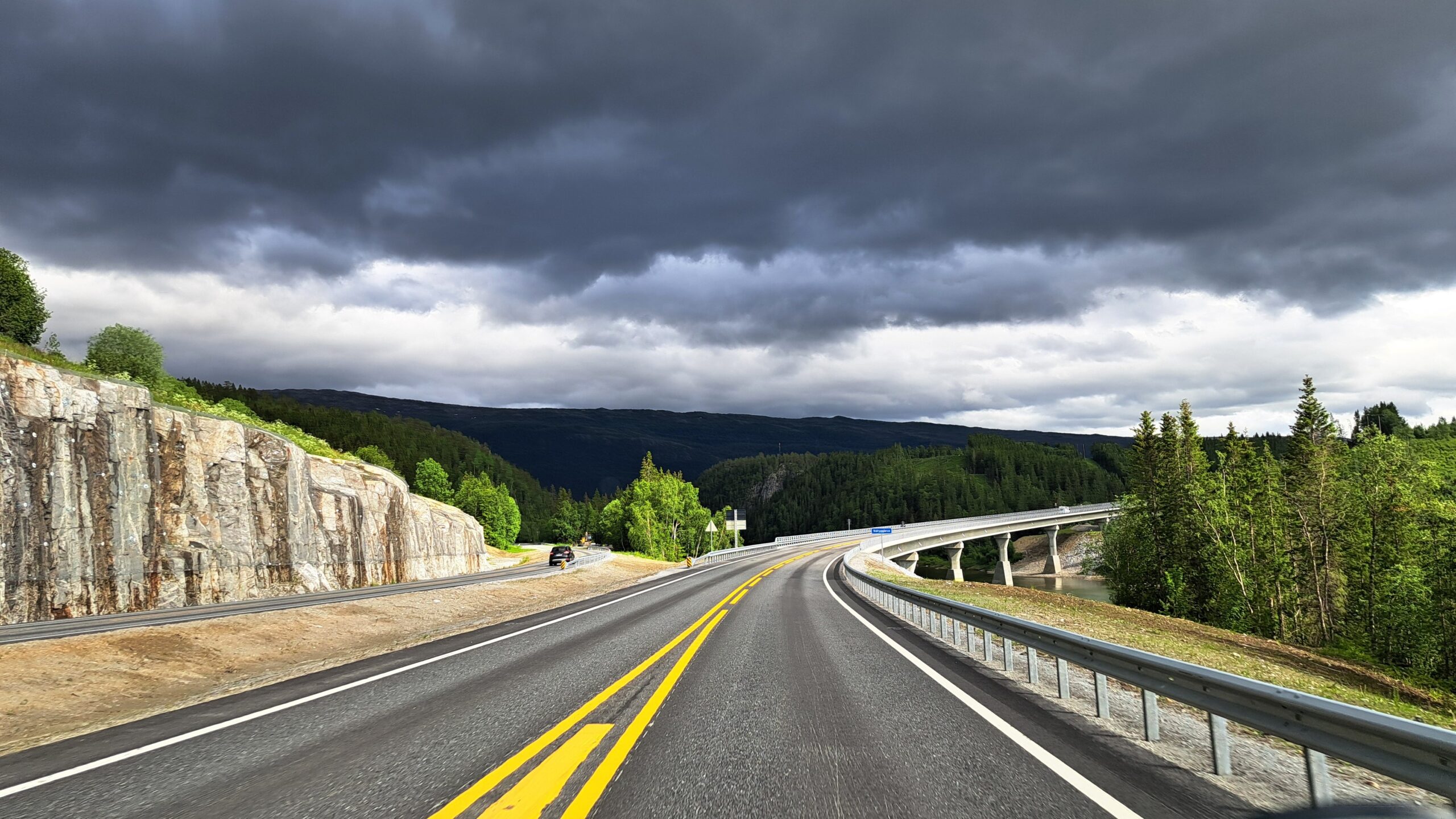 E6 in Norwegen durch bewaldete Berglandschaft unter dunklen Regenwolken